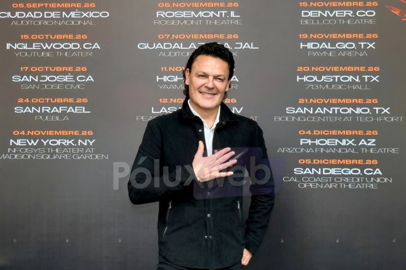 Fotografía en plano medio de Pedro Fernández sonriendo con una mano sobre el pecho y vistiendo chamarra negra y camisa blanca, posando frente a un banner promocional gris con las fechas y ciudades de la gira "Ave Fénix Two'r 2026".