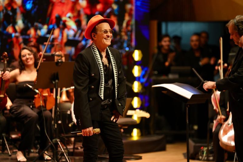 Fotografía en vivo del cantante Emmanuel sonriendo en el escenario con sombrero naranja y saco negro, micrófono en mano, rodeado por los músicos de una orquesta sinfónica con sus instrumentos de cuerda.