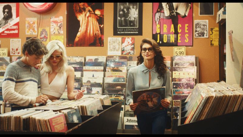 Fotograma promocional de la serie Fragmentos disponible en Disney Plus Tres jóvenes con vestimenta de época de los años 80 revisando discos de vinilo dentro de una tienda de música, con pósteres de artistas como Grace Jones, Def Leppard y Prince en la pared de fondo.
