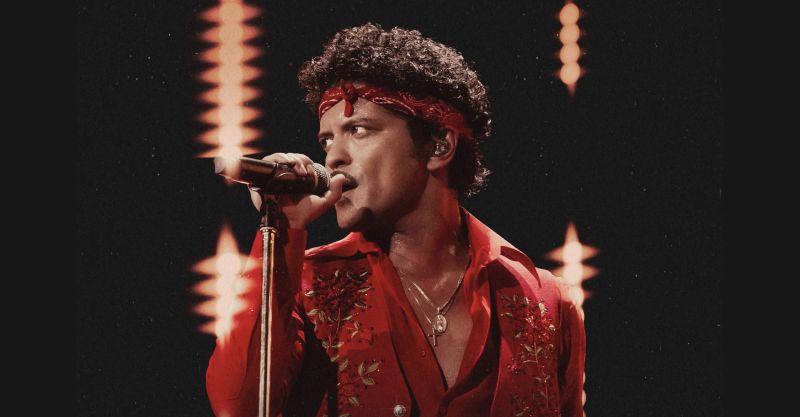 Bruno Mars anuncia quinta fecha de The Romantic Tour en el Estadio GNP Seguros Fotografía en primer plano de Bruno Mars cantando frente a un micrófono sobre el escenario con camisa roja desabotonada, paliacate rojo en la cabeza y luces de fondo.