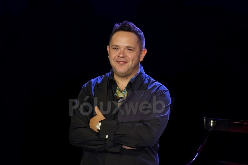 Rodrigo de la Cadena en la presentación del XI Festival Mundial del Bolero Fotografía de Rodrigo de la Cadena posando sonriente con los brazos cruzados junto a un piano de cola en un escenario con iluminación azul y fondo oscuro durante la presentación del evento.