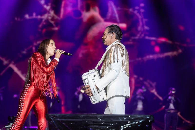 Presentación en vivo de Los Tigres del Norte Dos intérpretes en escena bajo iluminación púrpura y rosa, uno cantando y otro tocando acordeón, con pantalla gigante al fondo.