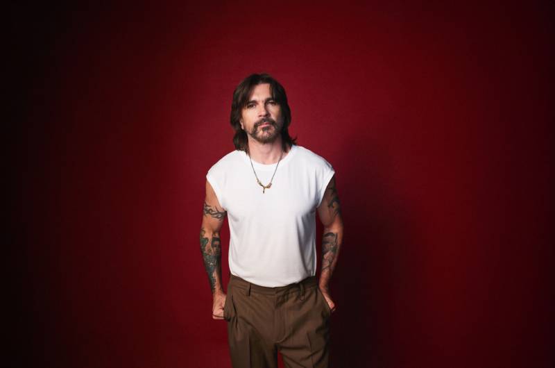 Retrato artístico de Juanes sobre fondo rojo Fotografía de una persona de pie frente a un fondo rojo, con cabello oscuro a los hombros, barba, camiseta sin mangas blanca y brazos tatuados, mirando directamente a la cámara.