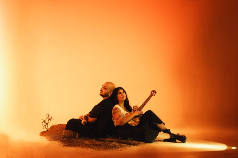 Dos artistas sentados espalda con espalda sobre un pequeño montículo de tierra, cada uno sosteniendo un instrumento musical, iluminados por un fondo cálido en tonos naranjas.