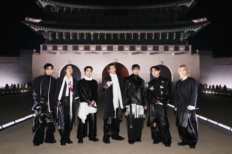 Siete integrantes posan frente a la puerta de Gwanghwamun iluminada de noche, vestidos con atuendos en blanco y negro y sosteniendo micrófonos.