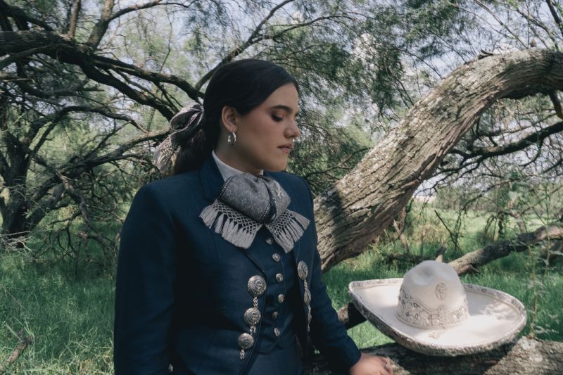 Camila Fernandez presenta su nuevo album Una Raya Mas Al Tigre Camila Fernández luce un traje charro azul marino con moño gris bordado y detalles plateados mientras mira hacia abajo en un entorno natural con un sombrero charro blanco a su lado. Archivo: General 2.jpg.