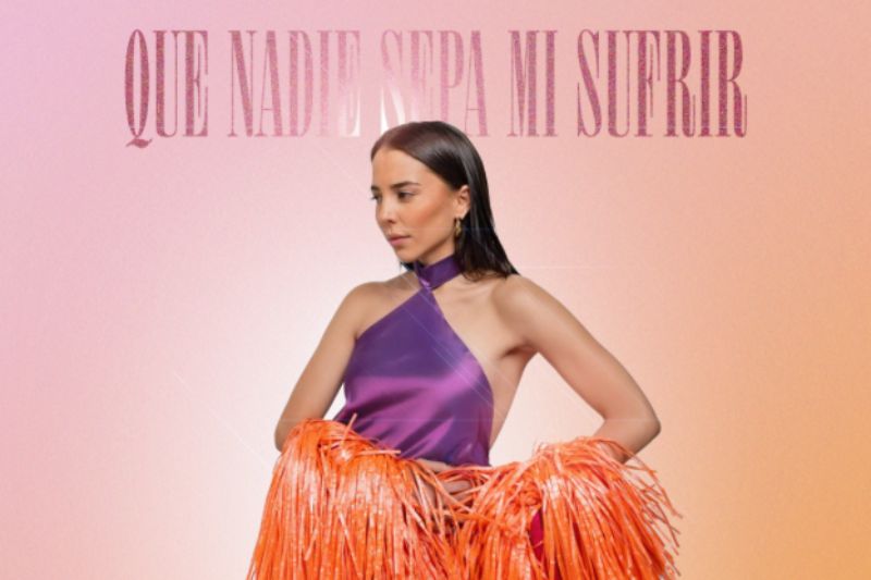 Majo Aguilar posa de perfil con la cabeza ligeramente inclinada luciendo un vestido morado de cuello halter y una capa naranja de tiras o flecos sobre un fondo con degradado rosa y naranja, con el titulo Que Nadie Sepa Mi Sufrir en la parte superior y su nombre en la parte inferior.