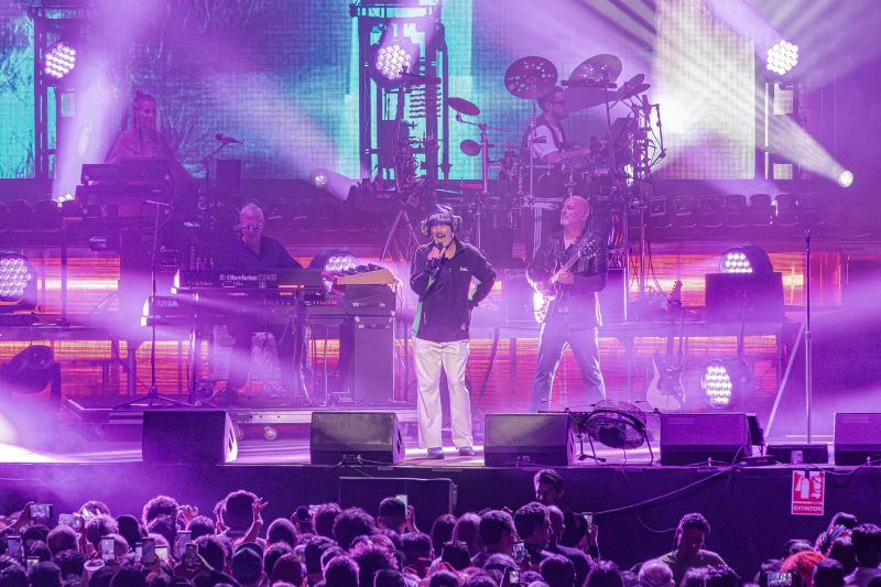 Jay Kay cantando al centro del escenario vistiendo sudadera y audífonos iluminados junto a los músicos de la banda bajo luces moradas.