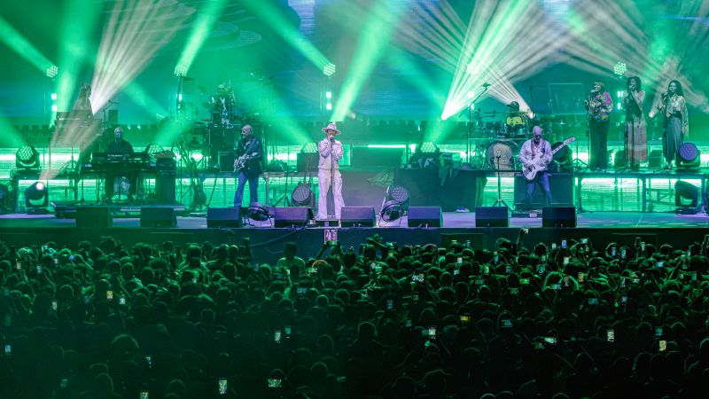 Vista frontal del escenario bañada en luces verdes con la alineación completa de Jamiroquai y la multitud con celulares encendidos.