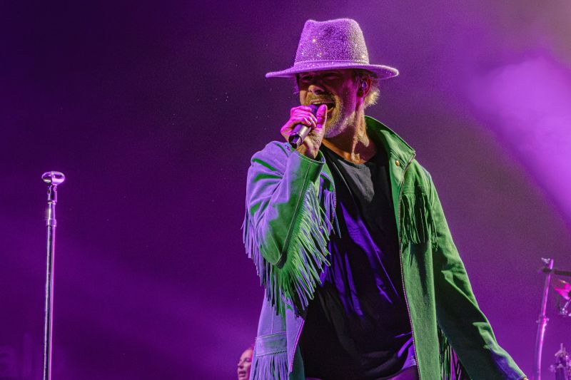 Primer plano de Jay Kay cantando con un sombrero brillante plateado y su chaqueta verde sobre fondo púrpura.