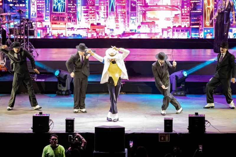El intérprete luciendo traje blanco, sombrero y leotardo dorado ejecutando la clásica inclinación de Smooth Criminal junto a sus bailarines vestidos de traje oscuro en el escenario.