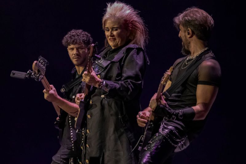 Jennifer Batten tocando la guitarra al centro flanqueada por el bajista y otro guitarrista del grupo musical sobre un fondo oscuro.