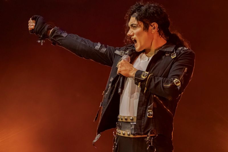 El cantante con el vestuario negro táctico de la era Bad alzando el brazo con el puño cerrado ante luces rojas de fondo.