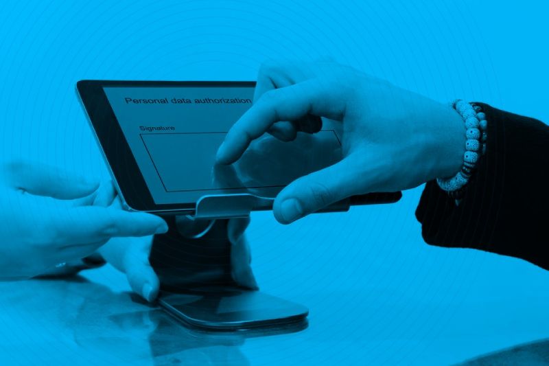 Firma electrónica digital en tablet con Sovos Sign y FAD® Fotografía en tono monocromático azul que muestra a una persona utilizando su dedo índice para firmar digitalmente un documento de autorización de datos personales en la pantalla de una tablet montada en un soporte sobre un escritorio.