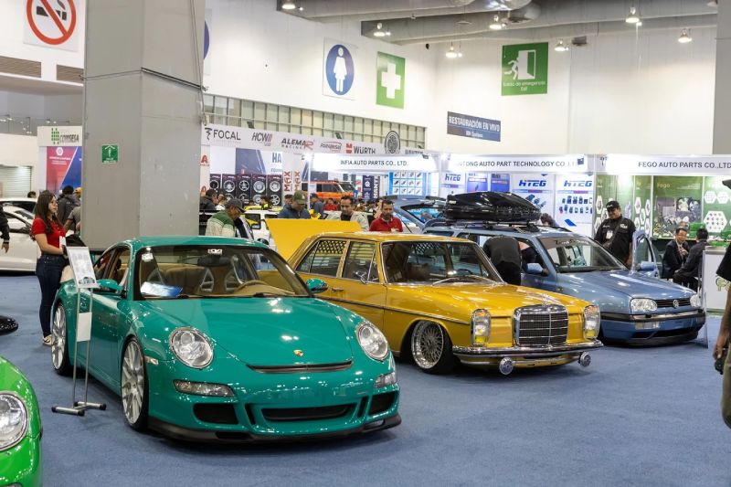 Fotografía del piso de exposición de INA PAACE Automechanika mostrando autos clásicos y deportivos modificados alineados, rodeados por estands de fabricantes de autopartes y asistentes.