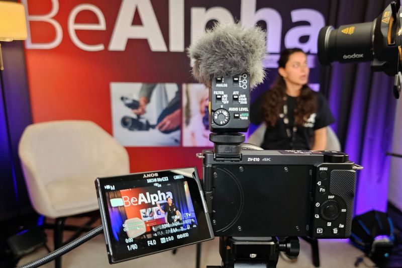 Cámara Sony ZV-E10 montada sobre un trípode con micrófono de cañón y pantalla abatible encendida con el logotipo Be Alpha, enfocando a una persona en un set de grabación.