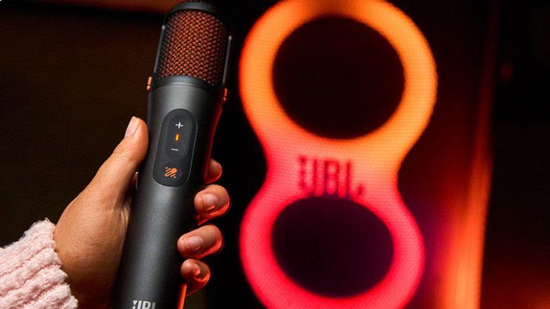 Micrófono inalámbrico JBL EasySing Mics con inteligencia artificial Un usuario sostiene con la mano un micrófono inalámbrico de color negro con la interfaz de controles iluminada en tono naranja. Al fondo se observa el panel de iluminación LED en forma de número ocho de un altavoz JBL PartyBox encendido en tonos rojos y naranjas.
