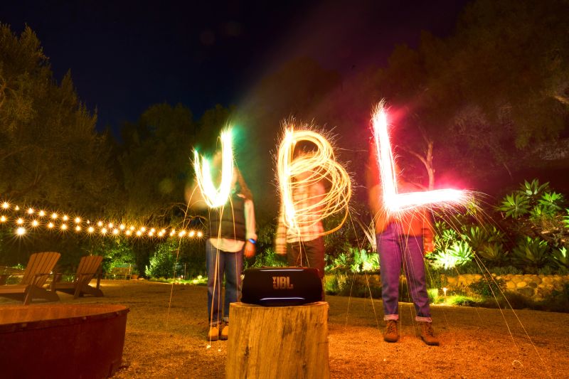 Tres personas sostienen luces de bengala que forman letras luminosas mientras una bocina JBL se muestra en primer plano sobre un tronco.