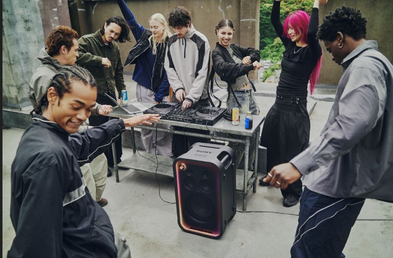 Grupo de jóvenes reunidos en una terraza al aire libre bailando alrededor de una mesa de DJ donde se encuentra una bocina de torre Sony ULT TOWER con luces encendidas alrededor de la rejilla.