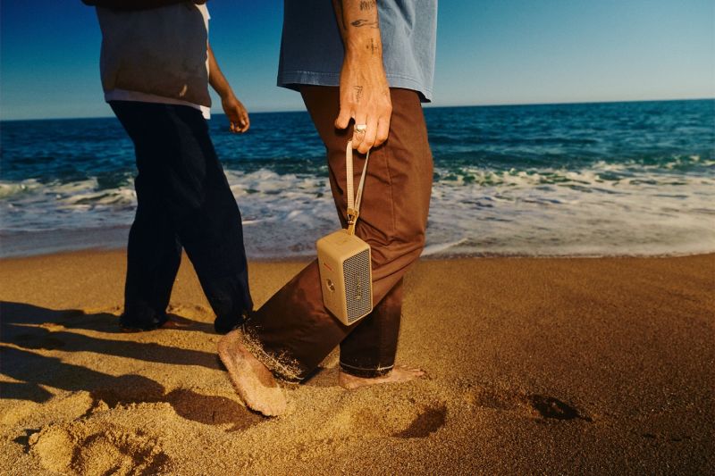 Una persona caminando descalza por la playa sosteniendo la bocina portátil Marshall Emberton III Brown mediante su correa, con el mar de fondo bajo la luz del sol.