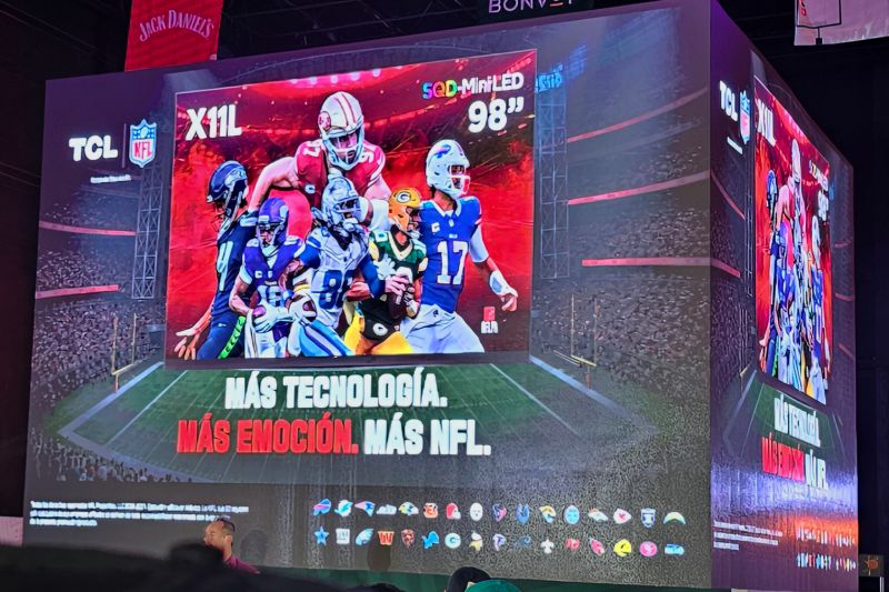 Una pantalla gigante cúbica en un evento de presentación donde se muestra la televisión TCL X11L de 98 pulgadas con tecnología SQD-MiniLED y gráficos de jugadores de la NFL sobre una cancha virtual.
