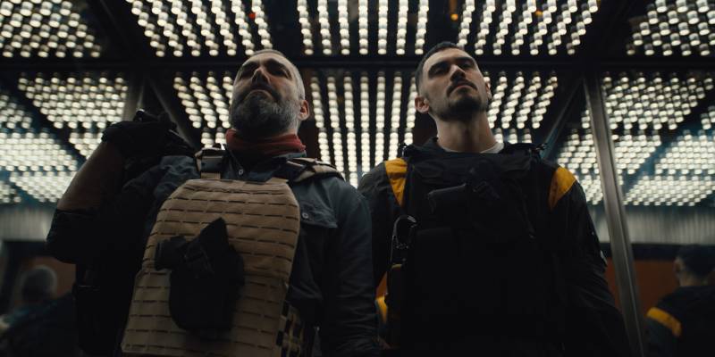 Dos personajes con equipo táctico se encuentran dentro de un elevador iluminado por paneles LED en el techo, creando un ambiente tenso y estilizado.