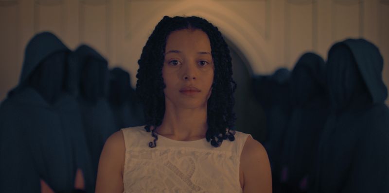 Ceremonia en sala oscura – Los Testamentos Una persona con vestido blanco se sitúa en primer plano, mientras varias figuras encapuchadas y vestidas de oscuro se alinean simétricamente en el fondo. El espacio interior tiene arcos y una iluminación tenue.