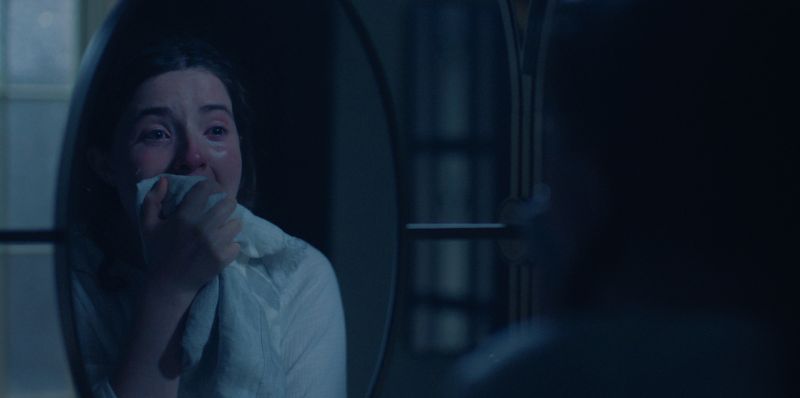 Persona mirando su reflejo en un espejo en una habitación tenue con luz azulada. Una persona observa su reflejo en un espejo dentro de una habitación oscura. Sostiene un paño cerca del rostro, iluminada por una luz azulada que acentúa la atmósfera introspectiva.