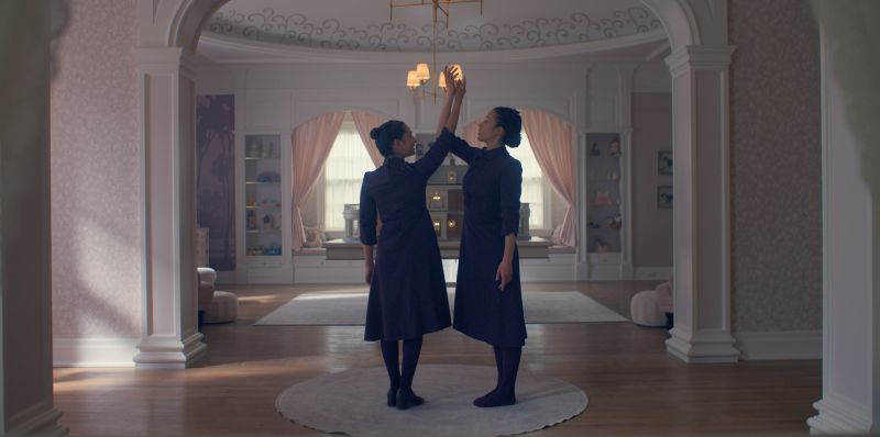 Ritual en sala ceremonial – Los Testamentos Dos personas vestidas con atuendos oscuros se enfrentan en una sala elegante, levantando un brazo en un gesto simétrico. El espacio está decorado con arcos, cortinas claras y mobiliario ordenado.