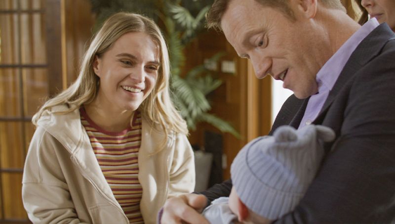 Fotografía promocional en plano medio de la película "Una Familia Perfecta" de Lifetime Movies. A la izquierda, una mujer joven rubia sonríe con calidez vistiendo una sudadera clara y playera a rayas. A la derecha, un hombre sonriente sostiene cuidadosamente en brazos a un bebé recién nacido envuelto con un gorrito tejido.