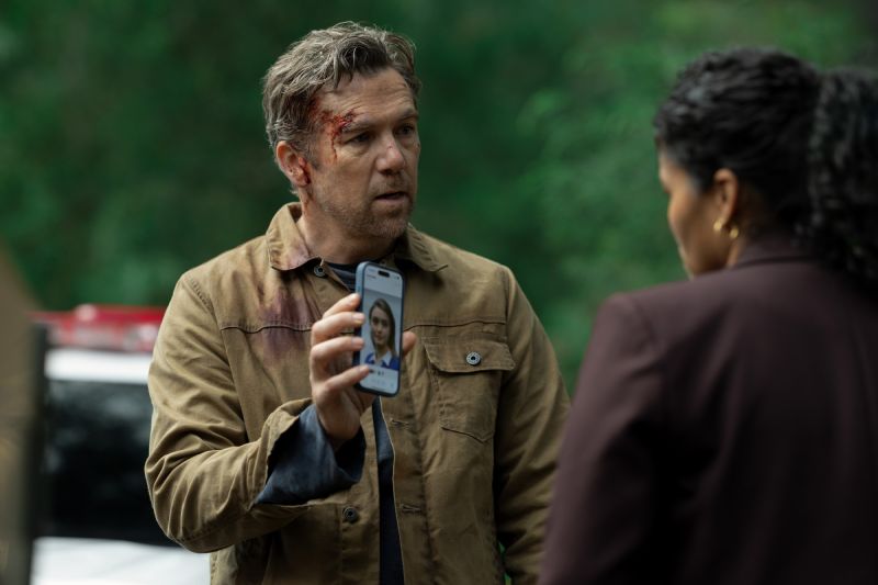 Patrick Brammall interpreta a Ian Ridley en la serie Vista por ultima vez de Apple TV Fotografía de escena en plano medio de Patrick Brammall personificando al detective Ian Ridley. El actor aparece con una herida con sangre en el lado derecho de la frente, vistiendo una chaqueta de pana café sobre una playera oscura. Sostiene frente a él un teléfono celular mostrando la fotografía de una joven en pantalla hacia otra persona de espaldas que viste un saco color guinda.