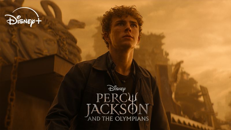 Fotografía promocional de la serie de Disney+ "Percy Jackson y los dioses del Olimpo". Walker Scobell interpreta a Percy Jackson en un plano medio, mirando hacia arriba con expresión seria contra un fondo cálido en tonos dorados que muestra una estatua clásica de Medusa y estructuras antiguas. En el centro inferior se muestra el logotipo oficial en inglés de la serie.