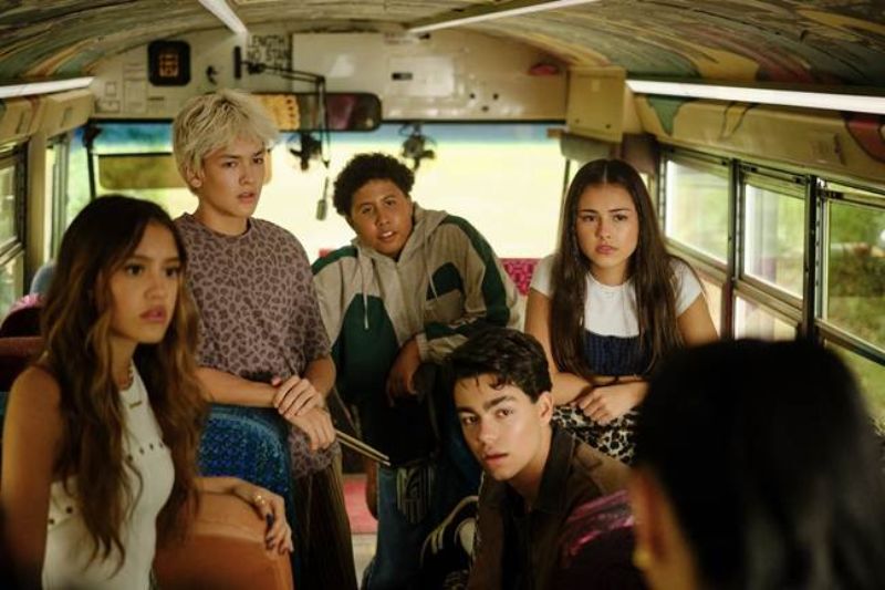 Nuevos campistas a bordo en Camp Rock 3 Seis jóvenes campistas sentados dentro de un autobús escolar retro conversando atentamente con miradas de sorpresa e intriga.