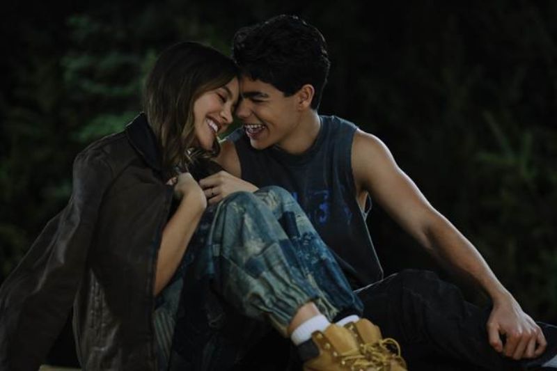 Romance juvenil en Camp Rock 3 Plano medio nocturno de dos jóvenes campistas sentados en el suelo sonriendo y compartiendo un momento íntimo con ropa invernal en el campamento.