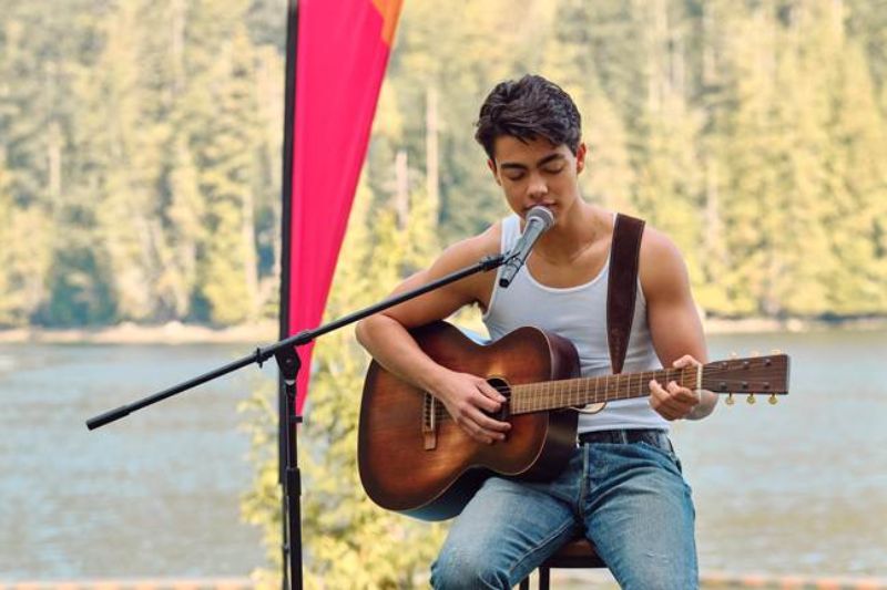 Presentación acústica frente al lago en Camp Rock 3 Un joven cantando concentrado frente a un micrófono mientras toca la guitarra acústica al aire libre con un lago y pinos de fondo.