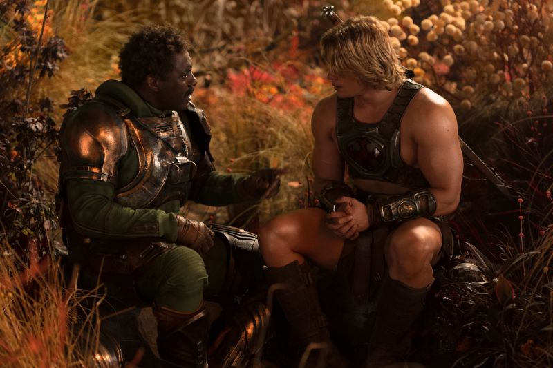 Fotografía fija de la película "Amos del Universo". En un entorno natural de vegetación alta y tonos otoñales, dos personajes conversan de perfil. A la izquierda, Duncan/Man-At-Arms, interpretado por Idris Elba con tez morena, viste una armadura metálica de tonos oscuros y bronce con acentos verdes en las mangas. A la derecha, el príncipe Adam/He-Man, interpretado por Nicholas Galitzine con cabello rubio semilargo, viste una armadura pectoral metálica con detalles ornamentales y sostiene sus manos entrelazadas mientras observa fijamente a su compañero.