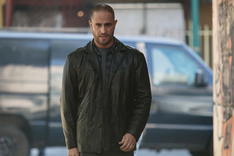 Plano medio del actor Michel Brown interpretando al detective Alejandro Falco. Viste una chaqueta de piel negra abierta sobre una playera azul marino con rayas horizontales oscuras. Luce el cabello rapado, barba corta y una expresión seria y concentrada mientras camina al aire libre en una calle urbana con una camioneta oscura estacionada de fondo.