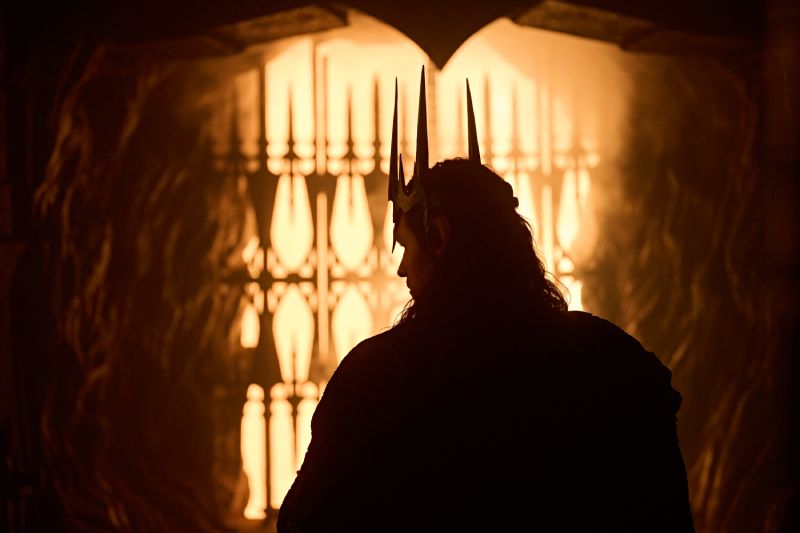 Fotografía cinematográfica a contraluz con tonos cálidos y dorados donde se observa la silueta oscura de espaldas del personaje Sauron, luciendo una corona de espinas o picos metálicos sobre su cabeza y una capa oscura. Al fondo, a través de un arco o portal con rejas de hierro talladas, se aprecia una luz brillante e intensa que evoca fuego y forjas.