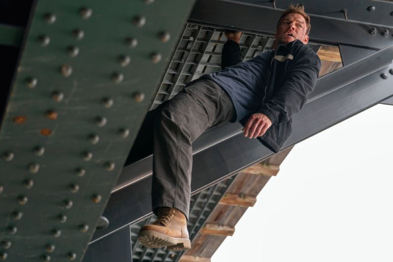 Fotografía de acción en plano medio con ángulo contrapicado. El actor Alan Ritchson interpretando a Jack Reacher cuelga de la estructura metálica inferior de un puente o viga industrial de acero con remaches. Viste una chamarra oscura con playera azul marino, pantalones cargo oscuros y botas de trabajo marrones; se aprecian raspones y heridas de combate en su cara y frente mientras mira hacia un lado con tono de urgencia.