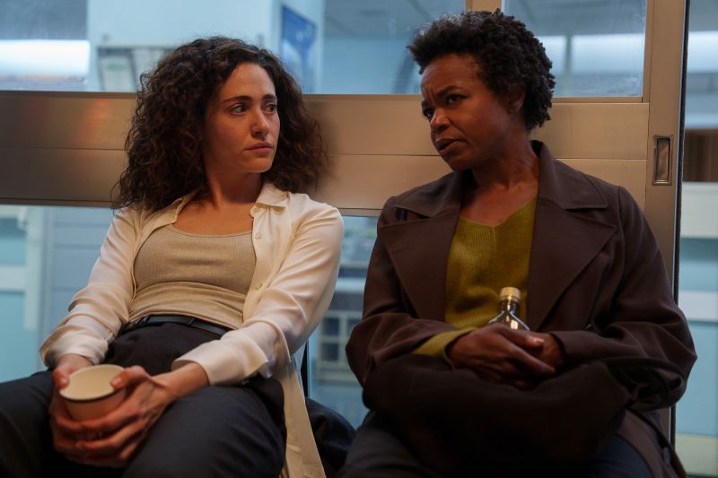 Emmy Rossum y Quincy Tyler Bernstine en la serie Furia Emmy Rossum y Quincy Tyler Bernstine sentadas juntas en un pasillo conversando serenamente mientras sostienen vasos de café en un momento de pausa dramática.