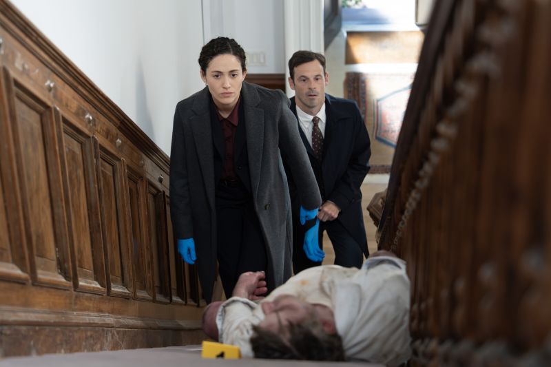Emmy Rossum en una escena de investigación en la serie Furia La agente Alice Black (Emmy Rossum) vistiendo abrigo gris y guantes de látex azules mientras sube cuidadosamente las escaleras de una residencia observando un cuerpo en el suelo durante una investigación policiaca.