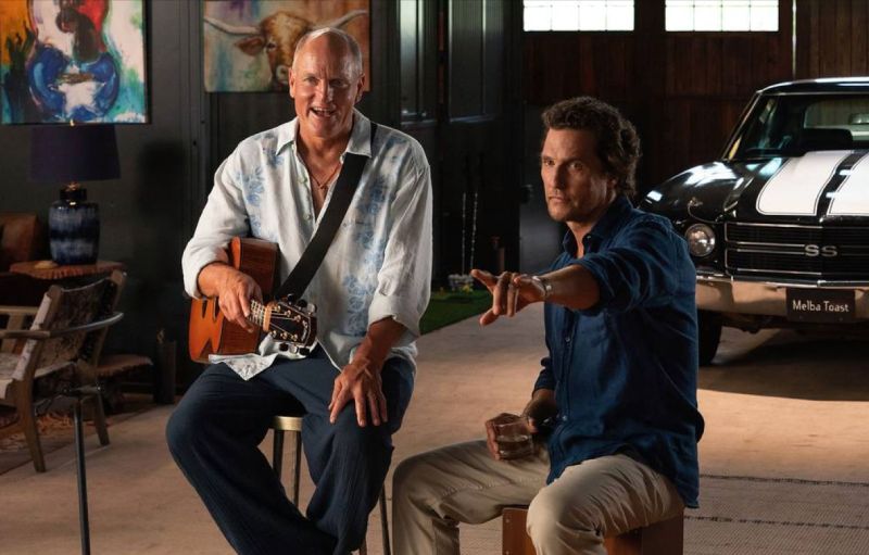 Dos personas conversan en un espacio tipo estudio o garaje, con una guitarra, un automóvil clásico y elementos decorativos alrededor.