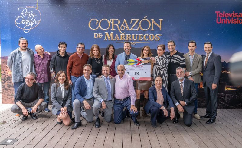 Grupo de personas posando frente a un fondo promocional con el título de una producción televisiva, sosteniendo una claqueta durante un evento de inicio de grabaciones.