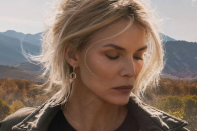 Persona de cabello claro recogido, con expresión introspectiva, situada frente a un paisaje de montañas y árboles otoñales iluminados por luz natural.