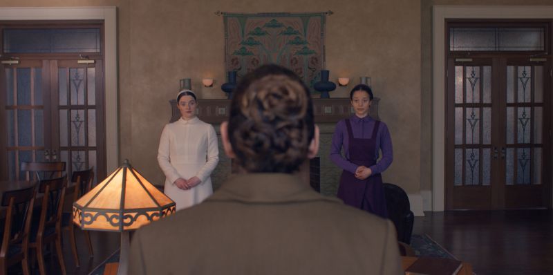 Los Testamentos – Renovación Temporada 2 Escena dramática en un salón formal donde dos personajes se enfrentan a una figura sentada, en un ambiente solemne y tenso.