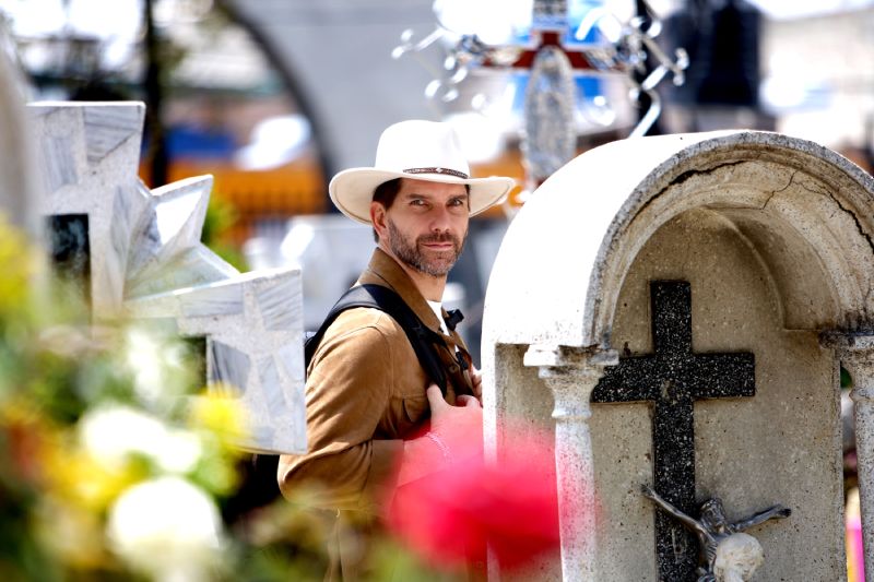 Arap Bethke conduce Mexico entre 2 Mundos en HISTORY Arap Bethke posa en primer plano luciendo un sombrero de ala ancha color beige y una chaqueta de gamuza marrón, sujetando las correas de una mochila. Al fondo, fuera de foco, se observan tumbas, cruces de mármol y flores en un panteón tradicional mexicano.