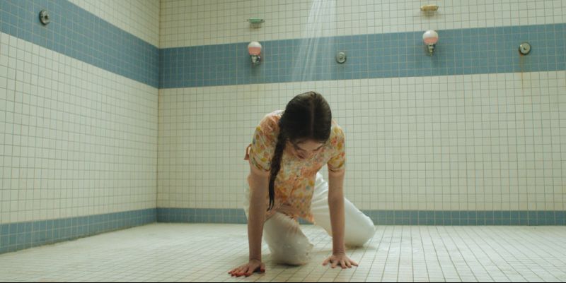 Summer H. Howell interpretando a Carrie arrodillada en el suelo de azulejos blancos de la regadera escolar bajo el chorro de agua.