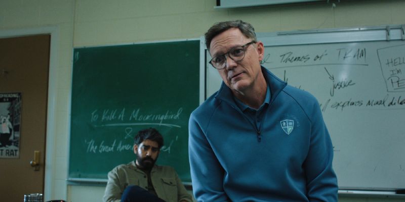 Un profesor en primer plano hablando en el salón de clases con un pizarrón verde al fondo y un estudiante sentado detrás.