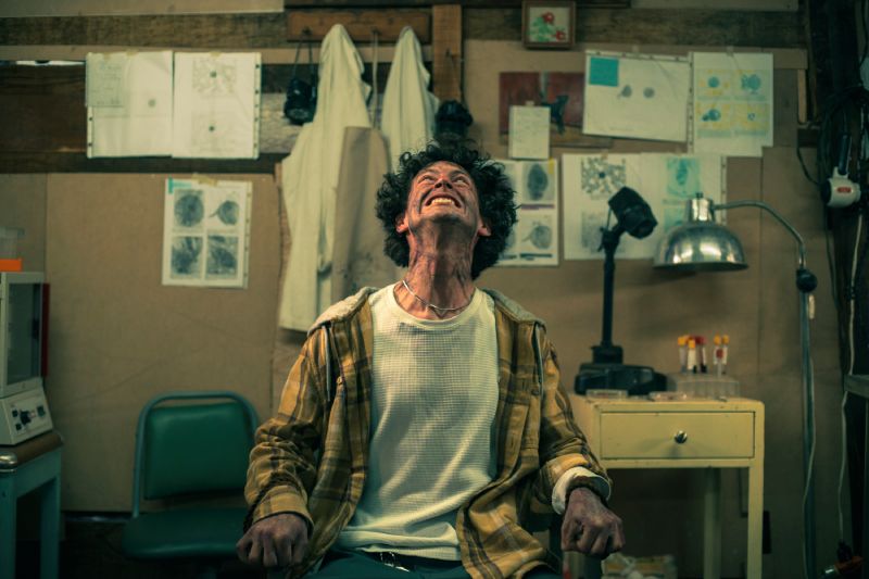 Un joven amarrado a una silla en un laboratorio improvisado gritando hacia el techo durante su transformación.