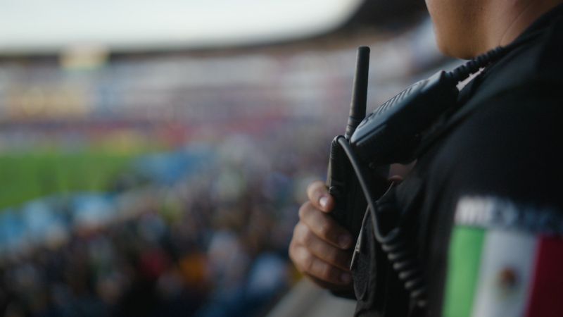 Primer plano del hombro y radio de un elemento de seguridad con el parche de la bandera de México observando hacia las tribunas.